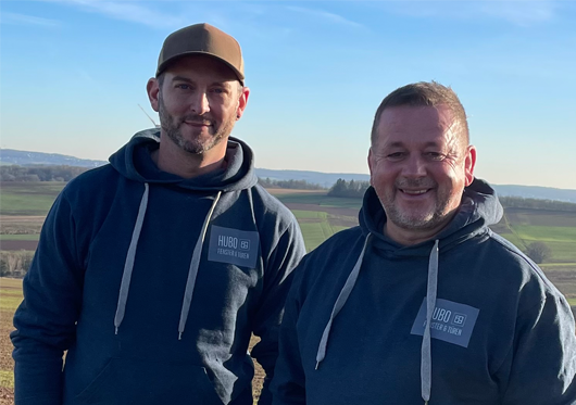 Die Geschäftsführer von HUBO Fenster & Türen - Zwei lächelnde Inhaber in Firmenkleidung vor einer ländlichen Kulisse in Lebach
